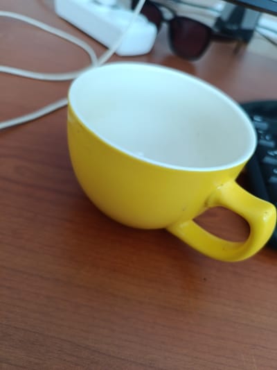 The image shows a close-up view of a yellow mug with a white interior resting on a wooden surface. The mug is positioned at an angle, with its handle clearly visible. In the background, out of focus, are a white object, likely an electrical adapter, and a charging cable. There are also a pair of dark sunglasses and a portion of a black keyboard visible. The lighting suggests an indoor setting, likely a desk. There are no people or animals present in the image. The scene appears to be a casual, everyday setting. The location of Baetov, Kyrgyzstan, is not discernible from the visual information provided in the image.