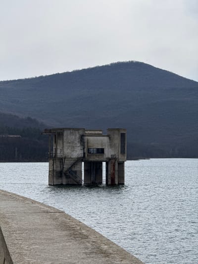 The image displays an abandoned, multi-level concrete structure, identified as a former pumping station at Pyasachnik Dam, near the city of Баня, Bulgaria. The structure is situated centrally within a large body of water, supported by concrete pillars partially submerged beneath the water's surface. Visible on the left side of the building is an external concrete staircase descending into the water. The brutalist-style concrete exhibits significant weathering, including dark stains and streaks of rust. Multiple window openings are present, some appearing to retain glass panes, others open. In the foreground, a rough, grey concrete walkway or dam edge extends diagonally from the lower left towards the center of the frame. The water is a consistent grey-blue, showing minor ripples. The background features a large, dark blue-grey mountain range covered in dense, dormant deciduous trees, set against an overcast, light-grey sky. No individuals, animals, or active elements are depicted in the scene.