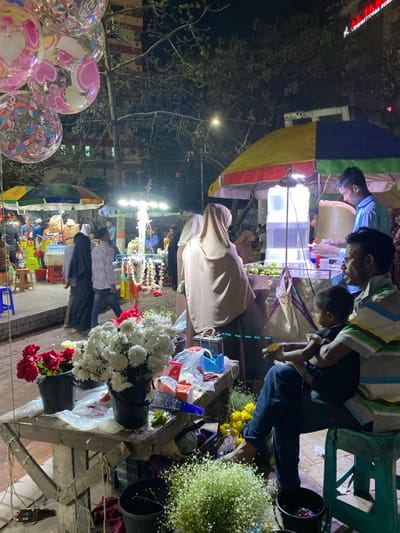 This image depicts a bustling night market scene in Narayanganj, Bangladesh. In the immediate foreground, to the upper left, several clear, spherical balloons adorned with pink and multicolored heart and swirl patterns are visible. Below them, a wooden vendor table holds a flower display. On the table, a black bucket contains red roses, and another black bucket holds numerous white chrysanthemum-like flowers. Various plastic bags, a blue tape dispenser, and small items are scattered on the table. Below the table, a dark bucket contains light green/white clustered flowers resembling baby's breath, and a dark bucket with yellow flowers is partially visible.

To the right, an adult male with a mustache, wearing a multi-striped short-sleeve shirt and dark trousers, sits on a green plastic stool. A young child, dressed in a dark shirt and shorts, sits on his lap, facing away from the viewer, holding a small yellow object.

In the midground, a paved pedestrian area is visible where several individuals are walking. These include a man in a light-colored shirt and two women dressed in dark abayas and hijabs. Further back, multiple street vendor stalls are set up under colorful umbrellas. One stall, brightly lit, features numerous garlands of flowers and beads hanging vertically. To the right, another vendor, a male in a light blue shirt, operates a drink stall under a yellow, red, and blue striped umbrella. He stands behind a counter with a large clear water dispenser and a large woven basket filled with a light-colored substance. Green fruits, possibly limes or lemons, are arranged on the counter. A woman in a light tan hijab and abaya stands with her back to the viewer, facing this drink stall.

The background shows dark tree branches, silhouetted against ambient light from streetlights and distant buildings. An illuminated sign for "INTERNATIONAL ACADEMY" is partially visible on one of the background buildings. The overall scene is illuminated by artificial lights from the stalls and streetlamps, indicating nighttime.