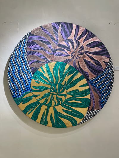The image shows a circular piece of artwork hanging on a light-colored wall, likely in Oaxaca City, Mexico. The artwork consists of two overlapping circles, one with a purple and brown spiral design and the other with a green and gold spiral design. The remaining space around the circles is filled with a geometric pattern of blue, white, and black triangles. A small white tag is visible at the top of the artwork. The wall is plain and slightly textured.