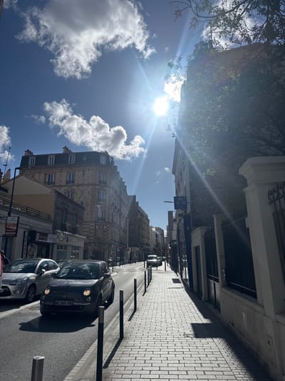The image depicts a bright, sunny daytime street scene in the city of Bois-Colombes, France. The perspective is from a paved sidewalk on the right, looking down a street lined with multi-story buildings.
The sky is bright blue with scattered white cumulus clouds. The sun, located in the upper right quadrant, emits a strong lens flare, casting long, sharp shadows towards the lower left across the sidewalk and street.
On the right, a light-colored, patterned brick sidewalk is bordered by a series of black, cylindrical bollards. Beyond the sidewalk, a stone wall topped with an ornate black wrought-iron fence partially obscures a building with darker brickwork and dense green foliage growing upwards. A blue sign with white text is visible on the building facade.
The street itself is paved with asphalt. A dark gray Fiat 500 (French license plate AZ-345-XO) is centrally positioned, facing away from the viewer, with its headlights illuminated. To its left, the front portion of a silver hatchback is visible, parked alongside the curb. Further down the street, another light-colored vehicle is partially visible.
On the left side of the street, a continuous row of multi-story buildings extends into the distance. The most prominent building in the mid-ground features a light-colored stone or brick facade with distinctive horizontal striped patterns on its lower levels and patterned stonework on upper floors, topped with a mansard roof and dormer windows. Shopfronts with French signage are visible at street level along this side. No pedestrians are distinctly visible.