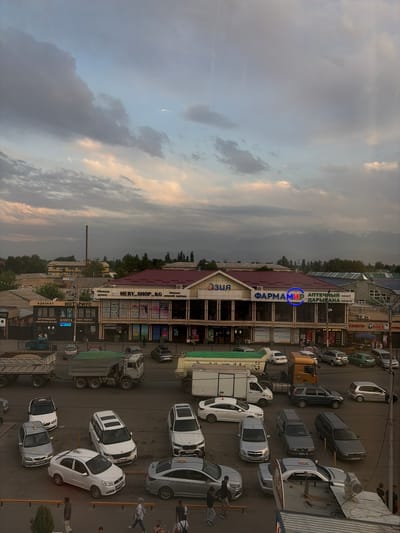The image shows a wide view of a street in Kant, Kyrgyzstan, with several buildings lining the road and a large number of vehicles. The sky is overcast with dramatic clouds, and mountains are visible in the distance.
In the foreground, several cars are parked and driving on the street, including sedans, SUVs, and a white pickup truck. A large white truck with a trailer is parked on the left side of the road.
The buildings in the middle ground appear to be a commercial strip with several storefronts. Signs indicate businesses such as a pharmacy ("ФАРМАМИР"), a clothing store ("MERY_SHOP.KG"), and a notary office ("НОТАРИУС"). There are people walking on the sidewalks in front of the shops.
In the background, more buildings and trees are visible, with the snow-capped mountains forming a backdrop. The overall impression is a bustling urban scene in a mountainous region.