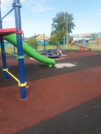The image shows a playground in Бородулиха ауылдық округі, Kazakhstan. The playground has a red and black rubber surface, a green slide, blue and yellow support structures, and a red seesaw. A child is sitting on a blue and green merry-go-round. In the background, there is a tree, a street light, and buildings. The sky is blue with some clouds.