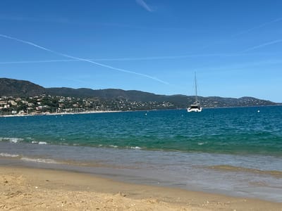 The image shows a serene beach scene in Le Lavandou, France. The foreground is dominated by a sandy beach with gentle waves lapping at the shore. The turquoise water stretches out towards a distant coastline, where a lush green hillside is dotted with houses and trees. A prominent sailboat with a white hull and tall mast is anchored in the bay, with a few other smaller boats visible in the distance. The sky above is a clear, vibrant blue, marked by wispy white contrails from airplanes. The overall atmosphere is peaceful and idyllic, characteristic of the French Riviera.