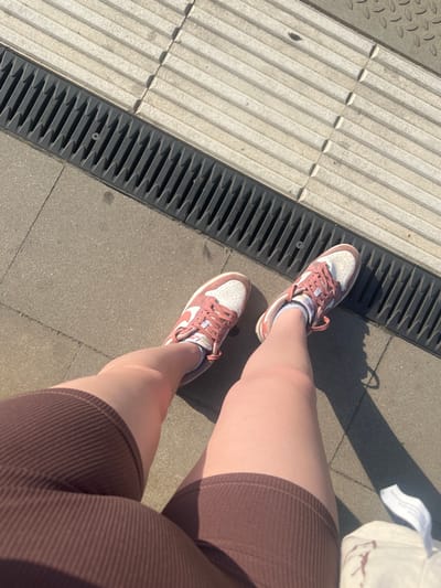 This media file shows a first-person perspective of a person sitting down outdoors on a sunny day. The subjects are the person's legs, wearing brown ribbed shorts and pink and white Nike sneakers. The setting appears to be a public walkway, with a textured, light-colored surface next to a dark, grated drain. The ground is paved with concrete tiles and cobblestones. Shadows indicate a bright, sunny day, likely in the afternoon. The person is seated, with their legs crossed and extended. The overall mood is casual and relaxed. No visible text is present in the image. The location context is Skawina, Poland.