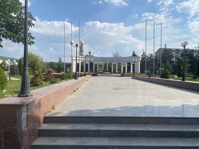 The image shows an outdoor plaza in Parkent, Uzbekistan, featuring a wide, paved walkway leading towards an arched colonnade. In the center of the plaza, a statue of a seated woman is visible within the colonnade. Several tall flagpoles stand in the background. The area is adorned with trees, manicured bushes, and flower beds. A man is standing on the left side of the image, near a lamppost and a granite-clad raised planter. The sky is blue with scattered white clouds, indicating daytime. The overall scene depicts a public monument or park.