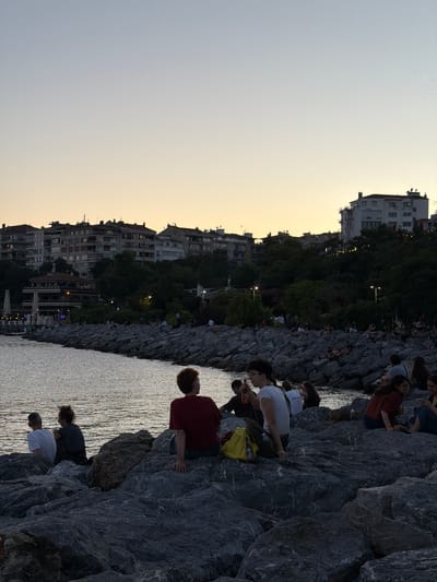 In Kadıköy, Turkey, people sit on large rocks by the water's edge at dusk. Buildings line the shore in the background, with trees and streetlights visible. The sky is a pale orange and blue, indicating the end of the day. Several individuals and small groups are seen relaxing, with some conversing and others looking out at the water. The water itself is calm, reflecting the soft light of the sky.