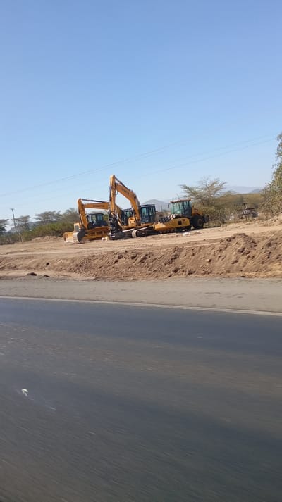 The image captures a daytime scene featuring several yellow heavy construction vehicles parked on a dirt area adjacent to an asphalt road.

In the foreground, a wide, dark gray asphalt road runs horizontally across the bottom third of the frame. The road surface appears somewhat blurred, suggesting the photograph was taken from a moving vehicle. Above the road, a raised dirt bank separates it from the area where the machinery is located.

On the dirt area, to the right of the dirt bank, a collection of construction equipment is parked. From left to right:
1.  A yellow track-type bulldozer or track loader is visible, with a substantial front blade (though mostly obscured by its own body) and track undercarriage. The brand name "SHANTUI" is clearly visible in black text on its side.
2.  Behind and slightly to the right of the bulldozer, two yellow hydraulic excavators are parked. The one on the left has its boom and stick partially raised, and its cabin is visible. The brand name "SHANTUI" is visible on its boom. The second excavator is partly obscured by the first one, but its yellow body and cabin are discernible. Both excavators appear to be tracked.
3.  Further to the right, a yellow wheel loader or possibly a large backhoe loader is partially visible, with large rear wheels prominent. Its front is mostly obscured.

The ground where the vehicles are parked is mostly barren dirt, showing signs of previous earthworks, with some small, dry shrubs and sparse vegetation scattered around.

In the background, beyond the construction vehicles, the landscape consists of more dry, scrubby trees and bushes, indicative of an arid or semi-arid environment. Distant hills or mountains can be seen under a vast, clear, bright blue sky. Thin power lines traverse the upper mid-ground of the image. The clear sky and bright conditions suggest it is daytime, likely mid-morning or afternoon. The vehicles appear to be stationary and not currently in operation, possibly parked at a staging area or a temporary storage site beside a road under construction or maintenance.