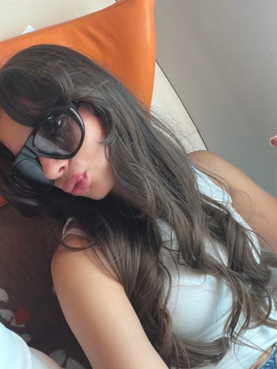 The image presents a close-up, tilted view of a person, likely female, reclining indoors. The subject's head is angled to the left, adorned with long, dark brown, wavy hair that flows across her body. She wears prominent, dark-lensed sunglasses that cover her eyes. Her lips are a focal point, colored with a pink or red lipstick and pursed in a distinct pout.
She is dressed in a light-colored, possibly white, ribbed top. A hint of blue denim fabric is visible in the upper right corner of the frame, suggesting she may be wearing jeans or shorts. To the left, an orange cushion or pillow adds a vibrant color element to the composition. The background largely consists of a plain, light gray or white surface, possibly a wall or ceiling, with a discernible seam. The person's right arm is partially visible in the lower right, resting on a dark surface with a subtle pattern, which could be a rug or upholstery.
The lighting appears soft and diffused, consistent with an indoor setting, creating a relaxed atmosphere. The image focuses on the subject's expression and style. No text is visible in the frame. The provided context indicates the location is Cagnes-sur-Mer, France.