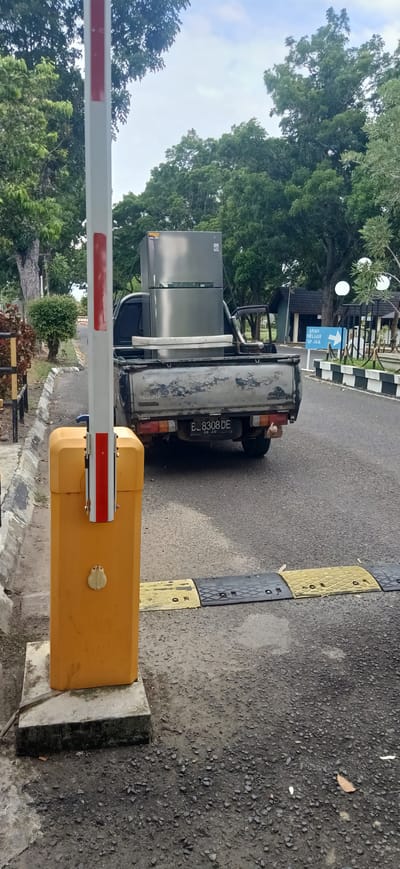 A silver LG refrigerator is being transported in the bed of a grey, older model Isuzu pickup truck. The truck is stopped at a road barrier. The barrier post is yellow with red and white stripes. The truck's license plate reads "B. 8308 DE" and has an expiration date of "06.20". Speed bumps are visible on the road in front of the truck. In the background, there are trees, a small building with a dark roof, and a blue sign with white text that reads "ARAH KELUAR SP JALAN" with an arrow pointing to the right. The scene appears to be outdoors during the daytime, with overcast skies suggesting a cloudy or potentially rainy day. The setting seems to be a road or driveway leading into a facility or area, possibly within Tambontunong, Indonesia, given the license plate format and tropical vegetation. There are no people visible in the image.