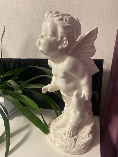 A white cherub statue is positioned on a white surface, possibly a shelf or table. The statue is facing to the left in profile, with its mouth puckered as if about to kiss. It has small wings on its back and stands on a textured base. To the left of the statue are green leaves of a plant, which are partially obscured by the statue. Behind the statue, there is a black reflective surface and a textured light-colored wall. To the right, a purple fabric curtain is visible. The overall lighting suggests an indoor setting, possibly a home. The image does not provide any information about a specific city or location, therefore no details about Старая Вичуга, Russia can be included.