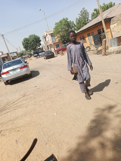 This image depicts an unpaved street scene in the city of Gama, Nigeria, under clear, sunny conditions. A young male, appearing to be an adolescent, walks towards the viewer's left in the foreground. He wears a light grey, long-sleeved tunic over grey trousers and dark sandals, carrying a light brown rectangular object in his right hand. His distinct shadow is cast to his right.
In the midground, a silver Honda Accord sedan is parked on the left side of the street, facing away from the viewer. Its rear license plate, with green text on a white background, is partially visible. Further down the street, a blue and white vehicle marked "POLICE" is present, accompanied by a dark sedan behind it and a red sedan parked on the right side. Several indistinct figures are visible near the police vehicle in the background.
Buildings, including both single and multi-story structures, line both sides of the street, featuring ochre and lighter-toned facades. Some buildings exhibit closed doors or windows. A motorcycle or bicycle is leaning against a building on the right. A tall street light pole with overhead power lines extends upwards in the midground. The ground consists of compacted dirt with visible tire tracks and foot impressions. Scattered green-leafed trees are visible in the background.