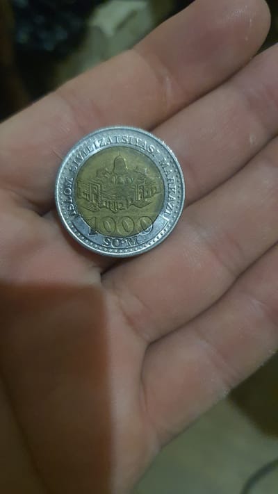 A close-up, overhead shot shows a person's hand holding a single coin. The coin has a silver-colored rim and a gold-colored inner circle. The inner circle features an illustration of a building with a dome, surrounded by smaller structures. Text is visible around the rim, reading "ISLOM SIVILIZATSIYASI MARKAZI," and the number "1000" is prominently displayed in the center, followed by "SOM." The coin is resting on the palm of a hand with visible wrinkles and skin texture. The background is blurred and appears to be an indoor setting.