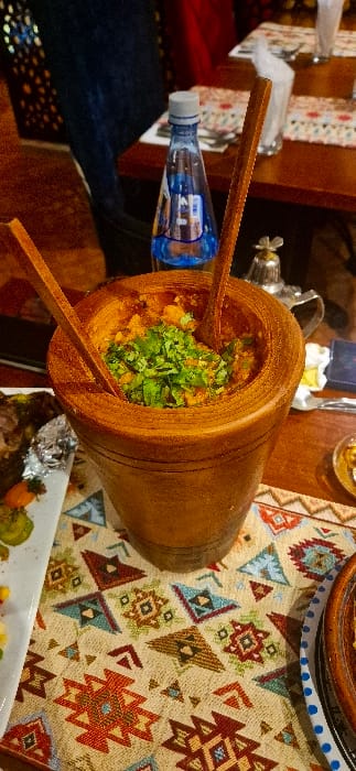 A wooden vessel, resembling a large mortar, is filled with a mashed food item and topped with fresh green herbs. Two long wooden spoons are inside the vessel. To the side is a small bottle of water. A colorful patterned tablecloth lies beneath the vessel and other objects. A white serving dish with food is partially visible at the bottom of the image.