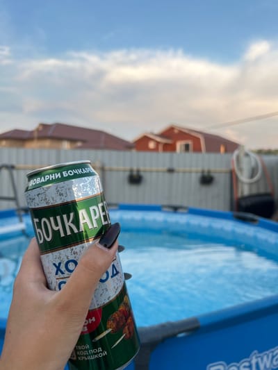 In the city of Solontsy, Russia, a person's hand with dark nail polish holds a green and silver can of beer in front of an outdoor swimming pool. The can has the text "БОЧКАРЕВ" and "ХОЛОД" on it, and features an image of meat skewers. The background shows a residential area with houses and a fence, under a cloudy sky. The pool appears to be filled with water and a portion of the "Bestway" logo is visible on its edge.