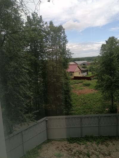 The image depicts a rural scene in the city of Komsomolsky, Russia. In the foreground, a corrugated metal fence with barbed wire runs horizontally across the frame, separating the viewer's perspective from the rest of the scene. Behind the fence, dense green foliage and trees dominate the left and right sides of the image, obscuring much of the background. In the mid-ground, a yellow house with a red roof is visible, along with a smaller adjacent structure and a red fence. Further in the background, several larger, industrial-looking buildings with grey roofs are scattered amongst more trees. The sky above is a mix of blue and white clouds. The overall impression is one of a somewhat overgrown, possibly residential or semi-rural area.