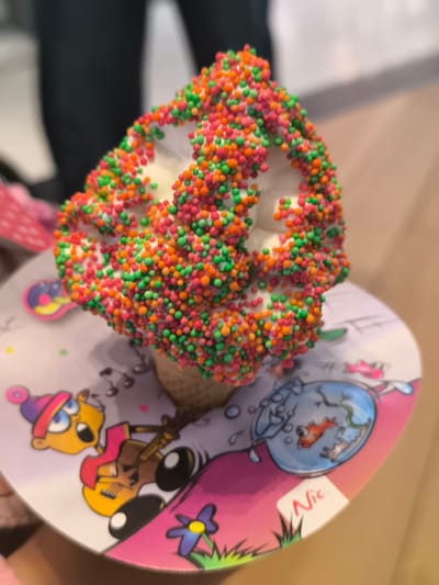 An ice cream cone rests on a small, round, disposable plate decorated with cartoon characters and a goldfish bowl image. The ice cream is vanilla, covered in colorful sprinkles of orange, green, and red. The cone is typical of ice cream cones. The scene is inside, as evidenced by the blurry background showing a person's legs and the interior flooring. On the plate is a sticker with the name "Nic" written on it. It is not possible to specifically identify the location in Leidschendam from the available evidence.