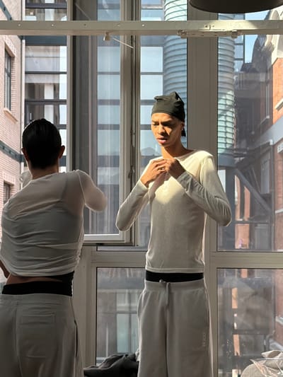 Saint-Denis, France. An indoor scene displays two individuals positioned near a large window. The individual on the left is seen from the back, in the process of adjusting a sheer, light-colored long-sleeve top, exposing a dark undergarment. They wear light-colored sweatpants with a black waistband. The individual on the right, facing slightly left, has dark skin, wears a black head covering, a light-colored long-sleeve top, and light-colored sweatpants with a black waistband; their hands are clasped at chest level. Strong, directional sunlight enters through the window, casting distinct shadows on the right individual. The background visible through the window reveals an urban setting with multi-story brick buildings, other windows, and a prominent ribbed cylindrical structure. A dark garment or object rests on the floor or window sill in the lower right foreground.