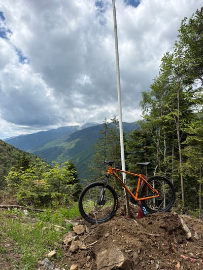 An orange mountain bike is positioned on a dirt ledge next to green foliage, set against a backdrop of mountainous terrain and a cloudy sky. A tall, white pole is visible in the foreground. The location is near Рилски манастир, Bulgaria.