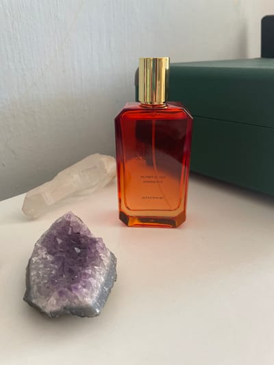 The image depicts a vanity scene, likely in a domestic setting, featuring a perfume bottle with a gradient of orange and red hues, topped with a gold cap. To the left of the perfume bottle are two crystals: a clear, elongated quartz crystal and a cluster of amethyst. In the background, a dark green box is visible. The lighting suggests an indoor environment, possibly near a window or under artificial light. The image does not contain any information or visual cues that would allow for its location to be determined as Kopeysk, Russia, or any specific city.