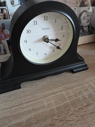 A black mantle clock with a white face is positioned on a light brown wooden surface. The clock has black hour and minute hands. The hour hand is pointing to approximately 3:30. The minute hand is pointing to the 7. In the background, out of focus, there are several framed pictures. One picture shows a man and a woman standing together, the woman is wearing a purple top and white pants, and the man is wearing a white t-shirt and dark shorts. Another picture is a black and white portrait of a young woman. The scene is indoors, on a table or shelf. There is no discernible information within the image to determine its location in the city of Brzeg, Poland.