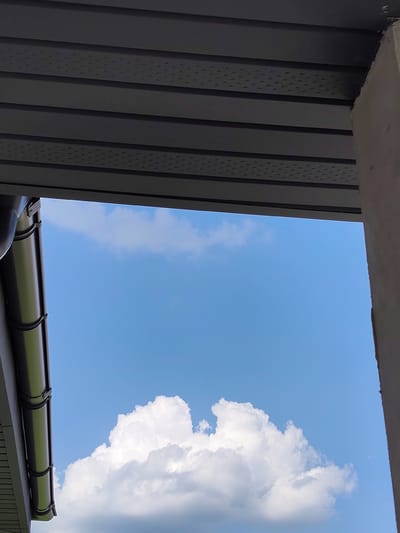 The image shows a view upwards towards a bright blue sky with scattered white clouds. In the foreground, a dark grey gutter system is visible on the left, attached to a dark grey overhang made of horizontal planks. On the right side of the image, a light grey concrete pillar or wall is present. The scene appears to be an outdoor setting, possibly a residential area, given the presence of architectural elements like the gutter and overhang. The overall impression is one of a clear day with some cloud cover. The location is identified as Skurygino, Russia. There are no people or animals visible in the image.