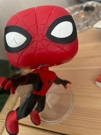 The image shows a close-up of a Spiderman action figure, likely a Funko Pop, in a red and black suit. The figure is positioned horizontally, with its large, red head with white eye lenses dominating the left side of the frame. The intricate red webbing pattern on the head is clearly visible. The figure's body is connected to its head by a metal post. The right side of the image shows the figure's torso, arms, and legs, also in red and black. The figure is supported by a clear plastic stand, which is mostly out of focus. The background consists of a light-colored wooden surface, possibly shelving, and some blurred objects in the upper left corner, one of which appears to be a clear plastic container or bottle. The lighting suggests an indoor setting, and there are no indications of weather or time of day. No text is visible in the image. The overall impression is that of a display of collectibles.