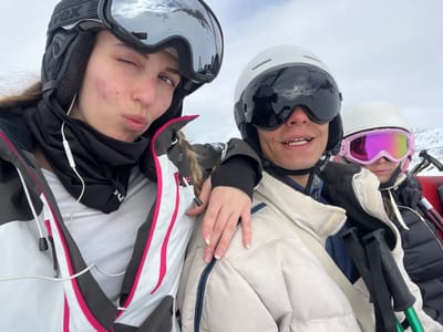 Three young individuals are captured in a selfie-style, close-up shot, appearing to be on a ski slope or lift. The original image is rotated 90 degrees clockwise, showing the subjects looking upwards towards the camera.
From left to right:
The leftmost person wears a white ski helmet and bright pink reflective goggles, with a dark jacket partially visible. Their face is mostly obscured by the goggles, but their mouth is visible.
The central person wears a white ski helmet and dark ski goggles, smiling broadly and showing their teeth. A hand with manicured nails (French tips) rests near their shoulder. They are wearing a light beige or tan ski jacket.
The rightmost person wears a black ski helmet and large mirrored ski goggles, with the brand "UVEX" visible on the goggle strap. They are making a playful face with slightly puckered lips and one eye partially closed. White wired earbuds are visible in their ears. They are wearing a distinctive white, black, and pink ski jacket.
Ski poles are partially visible on the far left of the image. The background is a bright, blurry white, indicative of a snowy environment or cloudy sky, consistent with a mountain resort setting like Val-d'Isère, France. The activity taking place is likely a break during skiing or snowboarding. The time of day is daylight, and the expressions suggest a cheerful and relaxed mood.