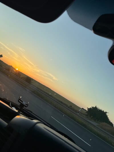 This is a photograph taken from inside a vehicle, looking out through the windshield at a sunset. The sun is low on the horizon, casting a warm orange and yellow glow across the sky, with some wispy clouds present. A road, likely a highway, stretches diagonally across the frame, with a white line marking the lanes. To the left of the road, a field of dry grass or crops can be seen, with a line of trees further in the distance. On the right side of the road, the edge of the vehicle's interior is visible, including a dark dashboard and a phone mount. The overall impression is one of a scenic drive during golden hour. There is no visible text in the image. The setting appears to be rural or semi-rural, likely in Germany, given the location context. The time of day is clearly sunset.