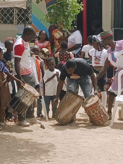 The image captures an outdoor gathering in Masvingo, Zimbabwe, centered on multiple individuals and drums under bright daylight. In the immediate foreground, the ground consists of light brown dirt or sand, showing distinct shadows.

Essential details:
*   **People:** Approximately 15-20 individuals are visible, encompassing adults and children of varying ages.
    *   In the foreground-left, a dark-skinned male adult, wearing a white short-sleeved shirt with red accents and blue jeans, holds a dark-colored drum with his left hand, looking towards the center-right.
    *   To his right, a dark-skinned male child, wearing a plain white short-sleeved t-shirt and blue jeans, stands with his hands clasped in front, looking downwards.
    *   Further right, a dark-skinned male adult, wearing a black short-sleeved t-shirt with a white "Ohio State" logo and ripped blue jeans, is bent forward, appearing to adjust or position a light brown drum that rests on the ground.
    *   On the far right, a dark-skinned individual, partially visible, wears a striped head covering, a white t-shirt with a monkey graphic, and a patterned skirt or fabric. Adjacent to them, another dark-skinned individual wears a multi-colored striped hat.
    *   Other individuals, including adults in patterned and plain clothing (some appearing traditional), and children, populate the midground and background.
*   **Objects:**
    *   Three large, cylindrical drums are prominent. One on the far left, held by the man in the white and red shirt, is dark-toned with lighter bands. The other two are on the ground near the bending man: one is light brown with vertical striations, and the other is decorated with a red, orange, and white abstract pattern.
    *   A small, light-colored object (possibly a pebble or debris) lies on the ground in the central foreground.
    *   A white plastic chair is partially visible on the far right.
*   **Environment:**
    *   The background features a light-colored building wall on the left with a window secured by a metal grille.
    *   A colorful mural, depicting abstract shapes in green, blue, and orange, is painted on a wall behind the central figures.
    *   Green foliage from a tree or bush extends into the upper center of the frame.
    *   A dark, recessed area or doorway is visible on the far right.
    *   The scene is illuminated by strong, overhead sunlight, casting defined shadows, indicating an outdoor daytime setting.
*   **Actions/Interactions:** The individuals appear engaged in a social gathering or performance context, with particular focus on the drums. The adults are either holding or attending to the drums, while children observe.
