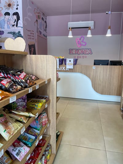 The image displays the interior of a retail store named "K-KOREA," located in Anápolis, Brazil. The perspective is from an aisle looking towards the cashier counter at the rear of the establishment.

To the left, a multi-tiered wooden shelving unit extends into the foreground, filled with various packaged goods, predominantly snacks, candies, and beverages, many bearing Korean script and identifiable branding. Visible price tags include "R$23.00" on some items. A large, unadorned, light-colored heart-shaped object is positioned on the top shelf.

In the midground and background, the cashier counter is a prominent feature, constructed with light wood paneling and a distinctive curved white front section. Two black computer monitors are positioned on the counter surface, alongside a white sign stating "CAIXA" (Cashier) and a promotional offer for "Lanche Brinde" (Free Snack) on purchases over R$50.00. Two purple plastic baskets containing items are also on the counter.

Above the counter, a neon sign illuminates the store's name, "K-KOREA," with a stylized pink and white 'K' logo, and Korean script "네 걸의 한국" below. The wall is horizontally divided into a light pink upper section and an off-white lower section. The pink upper wall features several cartoon-style illustrations of couples and text, including "Cause I'm falling slowly love with you." A white air conditioning unit is mounted on the wall above the counter. Three white pendant lights with wooden accents hang from the ceiling, providing illumination. A small, white security camera is affixed to the ceiling in the upper right quadrant of the image. The floor consists of large, light-colored square tiles. No individuals are present in the depicted scene.