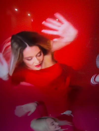 This image, captured from a digital display as indicated by the prominent screen door grid pattern and pixelation, depicts several individuals partially submerged in a large body of vibrant red and fuchsia-colored liquid. The scene is described as taking place in the city of Vladaya, Bulgaria.
In the upper central portion of the frame, a woman with light brown hair, wearing a red garment (possibly a dress or swimsuit) and red lipstick, is positioned with her face looking downwards and slightly to the right. Her right hand is raised clear of the liquid surface, with fingers splayed. Her left arm and hand are partially visible, submerged beneath the red liquid.
The liquid itself transitions in color, appearing as a deeper red in the upper half of the image and gradually shifting to a more fuchsia-pink hue in the lower half. Several bright white, indistinct reflections are visible on the red surface in the top section of the image.
Towards the lower left, a pale hand, likely belonging to another individual, is submerged in the fuchsia liquid. In the lower right quadrant, the face of another woman with blonde hair is visible, looking upwards with a slight smile. Her left arm is also discernible beneath the surface of the pink liquid. A partial view of a white and red striped arm accessory, possibly a bracelet or part of a garment, is visible on the far right edge of the frame.