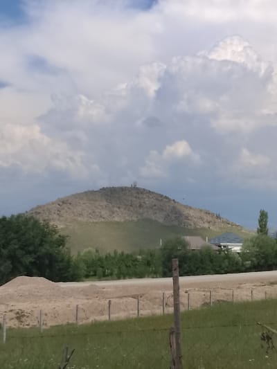 A broad, rocky hill dominates the center of the frame, with a prominent telecommunications tower at its summit. The hill is covered in loose rocks and sparse vegetation, with a green, grassy slope leading down towards the foreground. In the middle ground, several houses with modern architectural styles are nestled among trees. A dirt road runs horizontally across the mid-ground, separating the houses from a construction site with piles of sand and gravel in the immediate foreground. A simple wire fence with wooden posts encloses the construction area. The sky above is filled with large, fluffy white clouds against a light blue background. The overall scene depicts a rural or semi-rural environment on the outskirts of Kerben, Kyrgyzstan, with elements of nature, residential development, and ongoing construction.