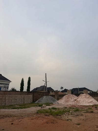 A residential area features single-family homes with dark rooftops. A pile of white sand and a pile of gray gravel suggest nearby construction. A tall power line stands prominently against the overcast sky. A brick wall borders the dirt road, along with three slender trees. The sky is overcast, creating flat lighting.