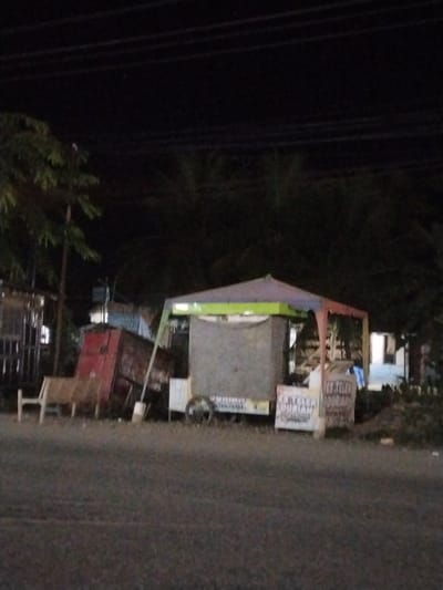 The image depicts a dimly lit outdoor scene at night, likely in Keudeteupinpunti, Indonesia. A small, covered food stall is visible in the center, with a multi-colored awning that is green, purple, and red. The stall has a grey fabric covering on its front, and signs with text that is mostly illegible due to the low light and image quality. One partially readable sign appears to say "ES TEH DURIAN." To the left of the stall, a red shipping container or large box is leaning against a wooden structure. In front of the container, a wooden bench is partially visible. Behind the stall, there are trees and some buildings with lights on. The ground in front of the stall appears to be a paved road or pathway. The overall atmosphere is quiet and somewhat deserted, typical of a street scene after dark.