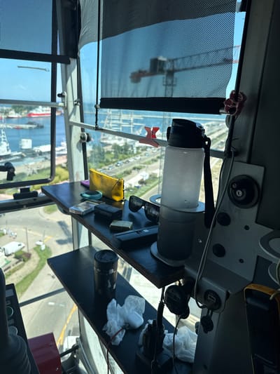 This image captures a close-up view of a cluttered shelf inside a crane operator's cab, offering a glimpse of the Gdynia harbor. The foreground is dominated by the interior of the cab, with various personal items resting on dark shelves.

On the upper shelf, a frosted water bottle with a black lid and strap stands prominently in a holder. Next to it are a pair of sunglasses, a small black rectangular object, a gold-colored lighter, and a pack of cigarettes. A yellow pouch and a small round brush are also visible.

The lower shelf holds a black travel mug, several crumpled white tissues, and what appears to be a pair of wireless earbuds. Electrical outlets and a portable charger are mounted on the right side of the cab.

Through the windows, a panoramic view of the Gdynia harbor unfolds. Several ships, including a red and white vessel, are docked in the blue water. The background reveals more of the harbor infrastructure, distant buildings, and a bright sky. The overall impression is one of a functional workspace, personalized with everyday objects, set against the backdrop of a busy port.