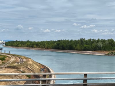 The image shows a wide, calm body of water, likely a canal, in Ottmarsheim, France. The water is a muted blue-green color and reflects the overcast sky, which is filled with scattered white clouds. Along the left bank of the canal, there is a steep, grassy embankment with a set of railroad tracks curving around it. Further up the bank, there are some industrial structures and what appears to be a building with a white facade. The right bank of the canal is lined with a dense forest of green trees. In the foreground, a metal railing is visible, suggesting the photo was taken from a bridge or elevated walkway. The overall scene is serene and industrial, hinting at the intersection of nature and infrastructure in the region.