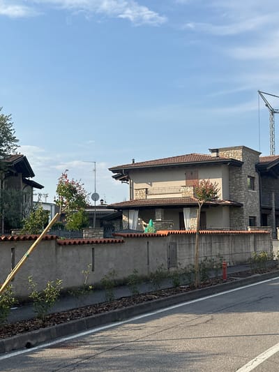 In the city of Rudiano, Italy, this image captures a residential street scene under a clear blue sky with scattered white clouds. A grey concrete wall with a red-tiled sawtooth-patterned coping runs horizontally across the foreground, separating the road from private properties. Several small, green-leafed shrubs are planted in a mulched strip between the wall and the asphalt road. A bright red fire hydrant is visible on the right side of the image.
Behind the wall, several modern houses are visible. The central house features a beige facade with stone accents and a red-tiled roof. It has a covered porch with white curtains and a second-story balcony with a black metal railing. A thin tree with pinkish-red blossoms stands in front of this house. To the left, another house with a darker stone facade and a steeply pitched roof is partially visible. A tall, thin wooden pole leans diagonally across the left side of the frame, positioned in front of the wall and the shrubs. In the background, a construction crane is visible against the sky, suggesting ongoing development. The road in the foreground is asphalt, marked with a white line.