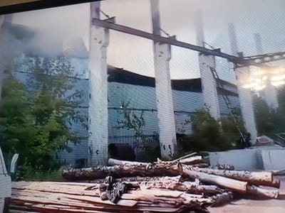 The image depicts the aftermath of a fire at an industrial site. Large concrete pillars and exposed metal beams, characteristic of a factory or warehouse structure, are visible. Some sections of the building appear damaged, with large open spaces and missing walls, suggesting structural compromise. In the foreground, a pile of cut logs and lumber is scattered across the ground, some of which appear charred. The ground itself is covered in what looks like rough terrain or debris.
To the left of the frame, dense green foliage from trees encroaches on the industrial scene, indicating proximity to nature or perhaps that the site has been disused for some time. There is a hazy, smoky atmosphere, particularly noticeable in the upper left portion of the image, which strongly suggests that a fire has recently occurred or is still smoldering. The sky is overcast or obscured by smoke, making it difficult to determine the time of day, though the lighting suggests it's daytime.
No people are clearly visible in the image, though there might be indistinct shapes in the background or obscured by the smoke. The overall mood conveyed is one of destruction and decay. The presence of logs could relate to the type of industry that was housed there (e.g., woodworking) or indicate materials that were stored or being processed on site. The location context provided is Vyalkovo, Russia, though no specific visual cues within the image directly confirm this. There is no visible text in the image.