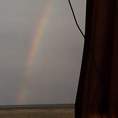 The image depicts a faint rainbow arcing across a grey, overcast sky. The rainbow is visible in the upper left quadrant of the frame. In the foreground, the right side of the image is dominated by a dark, vertical, textured object that appears to be a wooden post or structure. A thin black line, possibly a wire, runs diagonally from the top right towards the center of the image, near the rainbow. Below the sky, a horizontal line marks the horizon, with a muted, brownish landscape beneath it, suggesting a rural or undeveloped area. The overall impression is of a natural phenomenon observed under a somber sky, possibly from a sheltered vantage point. No elements in the image suggest a specific location within Sarul, Russia.