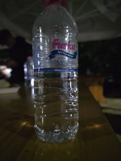 A horizontal close-up shows a 500 ml plastic bottle of Tuska spring water lying on its side on a wooden surface. The label is partially visible, revealing the brand name "Tuska" in pink and blue, along with the words "spring water" and "500 ml e" and the website address "www.lina07.com." The bottle appears mostly full and has some condensation droplets on its surface. The background is blurred, showing another bottle and indistinct shapes that might be people and objects. The lighting is dim. The location, Pripek, Bulgaria, is inferred from the water brand and the website printed on the label.