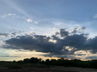 The image depicts a landscape scene featuring a dramatic sky over a treeline and a dirt ground. The sky is filled with a mixture of dark, heavy clouds and lighter, wispy clouds, with patches of soft, warm light suggesting either dawn or dusk. A dense line of dark green trees forms a horizon, with some lighter green foliage visible closer to the ground. The foreground consists of uneven, sandy or dirt ground with some piles of dark soil. The overall impression is one of natural beauty, with the sky being the dominant feature. There are no people or discernible structures visible in the image, and no actions or interactions are taking place. The image is likely a photograph taken in a rural or semi-rural setting. The prompt requests location information in Pisochyn, Ukraine, but this detail cannot be visually determined from the image itself.