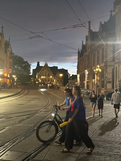 The image depicts a street scene in Ghent, Belgium, at dusk. The foreground features a bicycle with a yellow object attached to its frame, being ridden on tram tracks. Two women are in the process of crossing the street, with one pushing the bicycle. In the background, the historic architecture of Ghent is visible, illuminated by streetlights and the fading twilight. Several other individuals are walking on the cobblestone sidewalk. The sky is a deep blue, indicating the transition from day to night, and overhead wires for the tram system are prominent. The scene captures a typical evening in the city, with a blend of everyday activities and historical ambiance.