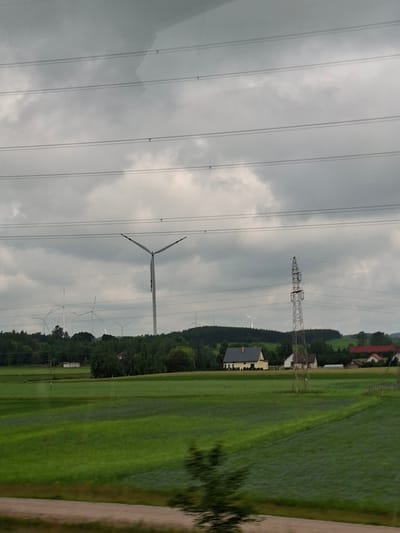 The image depicts a rural landscape under a cloudy sky. The main subjects are several wind turbines, some of which are in motion, suggesting they are generating electricity. The scene is a large, green field, possibly agricultural, with some darker patches that could indicate moisture or differing vegetation. In the background, a line of trees separates the field from a cluster of buildings, including houses with red and yellow roofs, and what appears to be a larger industrial structure, possibly a power pylon. The overall impression is one of a working landscape, focused on renewable energy production within a traditional rural setting. The weather appears overcast, typical of a cloudy day, and the time of day is indeterminate due to the lack of direct sunlight. No people are visible in the image, and there is no discernible text. The location context provided is Suwałki, Poland, which aligns with the presence of wind turbines and the rural architecture. The image is oriented vertically, making it a portrait-style photograph.