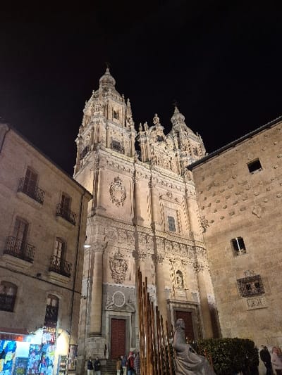 The image depicts a nighttime scene in Plaza de Anaya, Salamanca, Spain, dominated by the illuminated Baroque facade of the Pontifical University of Salamanca (Clerecía Church). The central building is a grand, multi-tiered stone structure featuring two ornate bell towers topped with spires and crosses. Its facade is richly detailed with columns, cornices, decorative carvings, crests, and niches containing statues. Two large red wooden doors serve as primary entrances at ground level, accessed via stone steps.

To the left, a multi-story stone building with several windows and wrought-iron balconies aligns with the university. Its ground floor houses a brightly lit commercial establishment with colorful merchandise displayed behind a glass front. To the right, the distinct facade of the Casa de las Conchas (House of Shells) is visible, characterized by numerous protruding, shell-shaped stone embellishments covering its surface. This building also features windows, including one with an ornate dark wrought-iron grille.

In the lower foreground and midground, a dark sculpture composed of multiple vertical, parallel elements stands centrally. To its right, partially obscured by the sculpture, a seated stone statue, identifiable as Fray Luis de León, is present. A low, green hedge runs along the base of the Casa de las Conchas.

Several indistinct human figures are visible at ground level. Approximately 5-7 individuals are gathered on the steps near the left entrance of the Pontifical University, while a few more are discernible on the far right at the base of the Casa de las Conchas. Their actions are limited to standing or walking. The sky above is dark and clear, indicating a night without cloud cover. Artificial warm-toned lighting illuminates the architectural subjects.