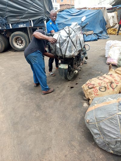 This outdoor image, captured in Satellite Town, Nigeria, depicts two men engaged in handling cargo on a motorcycle during daylight hours. In the foreground, a man wearing a black t-shirt, blue jeans, and brown shoes is bent over, actively securing or adjusting heavily wrapped items on the rear of a motorcycle. His arms show visible muscle definition, indicating effort. Behind him, another man in a blue polo shirt and sandals stands, observing or assisting with the task.
The motorcycle is heavily laden with objects tightly wrapped in clear plastic and secured with tape or rope, suggesting a substantial amount of goods being prepared for transport. The license plate on the motorcycle clearly displays "LAGOS SW-158GR." Dark liquid stains are visible on the asphalt ground near the motorcycle's rear wheel.
In the background, a large blue truck is parked, with the word "ONLY" partially visible on its side. Large truck tires are prominent. The scene also features various sacks and bags on the ground. One distinct burlap sack is marked with "FISH," "COD," "30 KILOS NET," "PRODUCT OF NORWAY," and "EFTA," indicating its contents are imported fish. Other goods are covered with large blue tarpaulins, consistent with a busy loading or commercial area. The overall impression is one of a logistical operation or market activity.