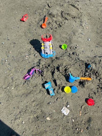 This media file showcases a collection of children's beach toys scattered on wet, dark sand. The toys include a red bucket, a blue and yellow sand shovel, a green bucket, a red and grey plastic vehicle, a blue truck, pink swimming goggles, and other small plastic pieces. Some of the toys appear to be covered in sand. The scene is outdoors, likely at a beach, given the sand and the presence of beach toys. The lighting suggests it is daytime, with strong sunlight casting shadows. The ground is predominantly sand with small pebbles and shells. The overall impression is one of a recent play session at the beach. There is no visible text in the image. The location context is Germasogeia Municipality, Cyprus, which is consistent with a beach setting.