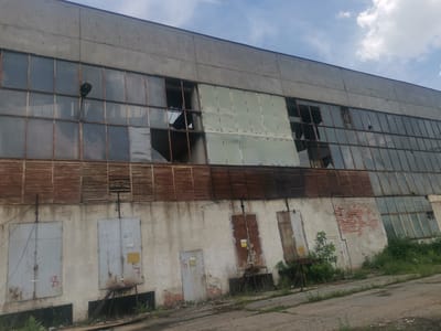 This image captures a dilapidated, multi-story industrial building, likely an abandoned factory or warehouse, under a partially cloudy daytime sky. The overall scene conveys a strong sense of neglect and decay.
The upper section of the building features a large, grey concrete facade. Below this, a vast expanse of windows runs along the entire length of the structure. Many of these windows are broken, with shattered panes, missing glass, and bent frames. Some sections appear to be boarded up or covered with translucent material, further contributing to the building's disused appearance. A black, large light fixture or street lamp is visible attached to the window frame in one section. Below the windows, a band of rusty, horizontal louver-like structures, possibly remnants of an old ventilation system, stretches across the building.
The lower half of the building's wall is concrete, showing signs of peeling paint, grime, and wear. Several metal doors are haphazardly placed along this section. Two large, light grey metal doors are prominent on the left, with "8" and "7" spray-painted in red on them, respectively. Smaller, unreadable labels and stickers are also present, with one partly legible as "ОБЪЕКТ..." on the second large door. Further to the right, a smaller grey door is marked with a red "6" and a yellow sign with "ПРЕКЪСНАТ..." (likely indicating "interrupted" or "disconnected"). Another heavily rusted, brown metal door is visible, next to a section of exposed red brickwork in the wall.
The ground in front of the building is a cracked, uneven surface, partially covered with dirt and overgrown with weeds and small bushes, particularly dense on the right side. The clear sky suggests fair weather, contrasting with the building's dilapidated state. No people or activities are present, emphasizing the building's abandonment. The location context of Zhivkovo, Bulgaria, suggests this is an industrial ruin in the region.