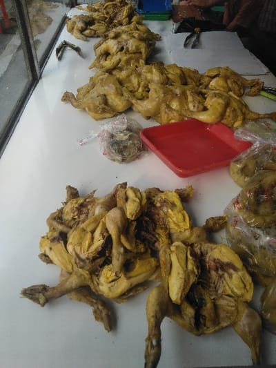 The image displays a food stall or market counter located in Cipanas, Indonesia. Numerous cooked chickens, exhibiting a prominent yellowish coloration, are laid out across a white countertop. The chickens are presented both whole and split open, with internal cavities visible on several specimens. They are arranged in multiple rows and piles. On the right side of the counter, an empty red plastic tray is present, alongside two clear plastic bags containing smaller, unidentifiable food items. In the upper background, a pair of tongs with black handles, a blue plastic container, and a green plastic container are visible. A person's arm, clothed in a brown-patterned fabric, is partially discernible in the upper right. The left side of the scene is bordered by a clear glass panel, likely part of a display case.