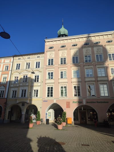 The image captures a view of the Max-Josefs-Platz in Rosenheim, Germany. The scene is dominated by the facades of traditional European buildings with pastel-colored walls and white decorative elements. The building on the right, painted in a light pink hue, features a prominent green dome topping its structure, visible against a clear blue sky. This building also displays an inscription that reads "Karl Jäger Bayerisches Trachtenhaus." Arched walkways form the ground level of these structures, revealing shops and storefronts. Paved cobblestone forms the ground surface. Shadows cast by the buildings and possibly trees are visible on the ground, indicating the time of day. Several potted plants are placed near the walkways, and a few individuals are present in the scene, some appearing to be shoppers or passersby.